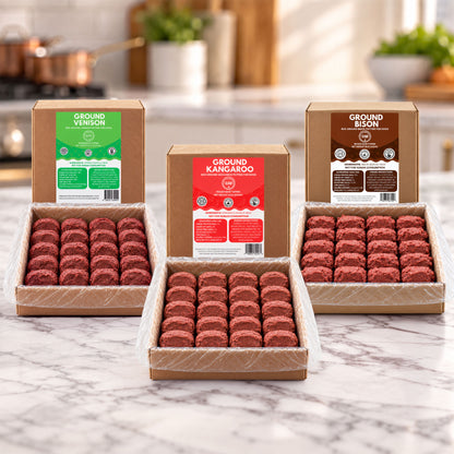 Three open cardboard boxes of ground meat patties arranged on a marble countertop in a kitchen setting. The center box is labeled Ground Kangaroo, flanked by Ground Venison and Ground Bison boxes behind it.
